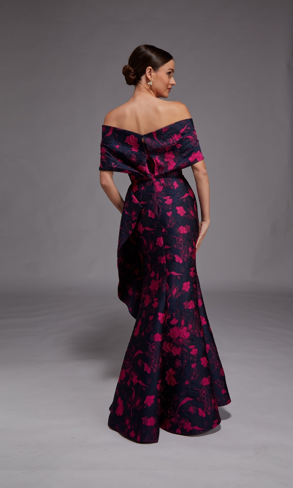 A woman faces away, wearing the Alyce 27832 Long Formal Dress—an off-the-shoulder, floor-length gown in dark floral jacquard with pink blooms—posed before a plain gray background.